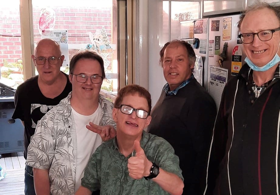 The birthday boy Peter is front and centre with two of his male friends standing on either side of him. Everyone is smiling and looking at the camera while Peter presents the thumbs up hand gesture. They appear to be standing in the kitchen as you can see a fridge in the background.