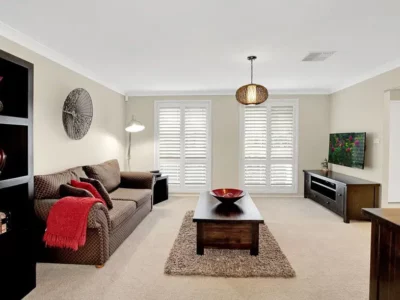 Living room with cream carpet, brown rug, brown sofa, two wooden side tables and a bookshelf. A television is mounted to the wall.