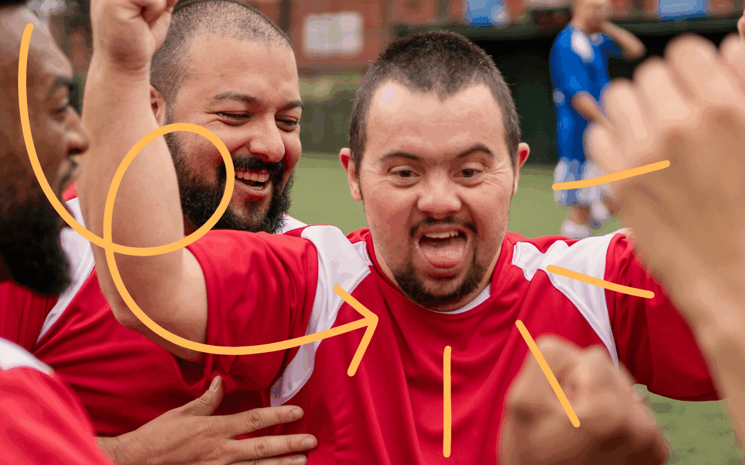 A man with a disability grins and holds up his arms in excitement. He is wearing a soccer uniform and is surrounded by happy teammates congratulating him.