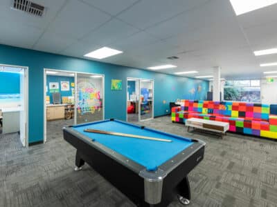Large room featuring a pool table and a colourful sofa. Smaller activity rooms can be seen in the background.